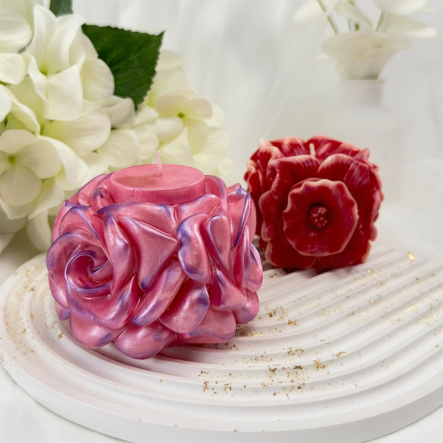 Zwei handgefertigte dekorative Kerzen in Blütenform – eine schimmernde rosa Rose und eine dunkelrote Anemone – auf einem weißen gewellten Tablett.