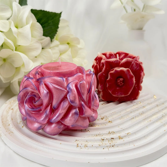 Zwei handgefertigte dekorative Kerzen in Blütenform – eine schimmernde rosa Rose und eine dunkelrote Anemone – auf einem weißen gewellten Tablett.