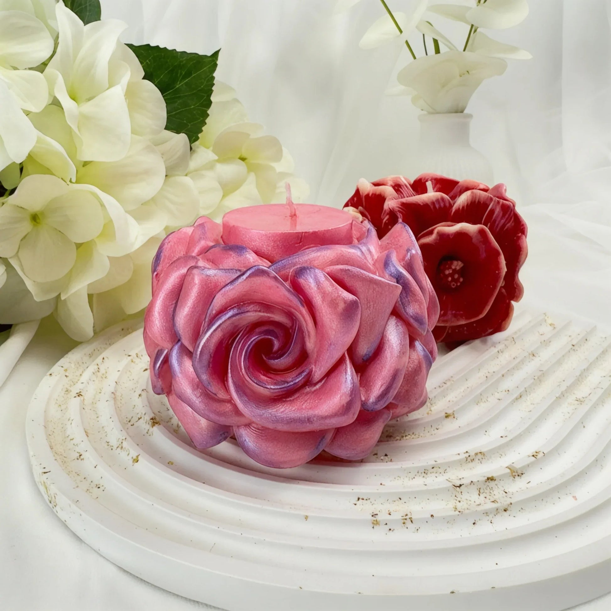 Zwei handgefertigte dekorative Kerzen in Blütenform – eine schimmernde rosa Rose und eine dunkelrote Anemone – auf einem weißen gewellten Tablett.