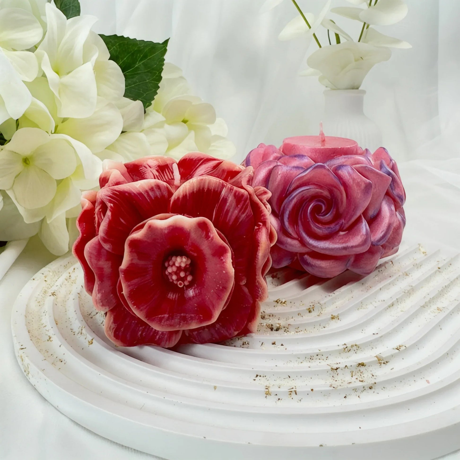 Zwei handgefertigte Blütenkerzen – eine dunkelrote Anemone im Vordergrund und eine schimmernde rosa Rose dahinter – auf einem weißen gewellten Tablett.