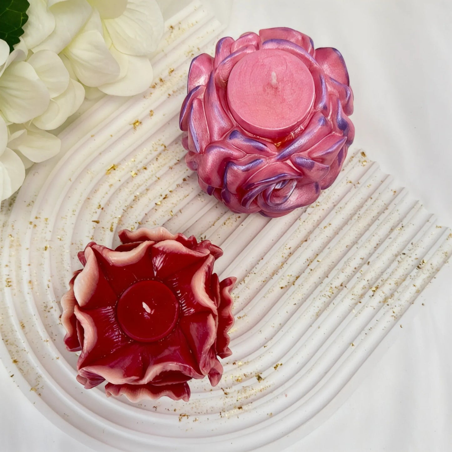 Draufsicht auf zwei handgefertigte Kerzen in Blütenform – eine schimmernde rosa Rose und eine dunkelrote Blüte – auf einem weißen gewellten Dekotablett.