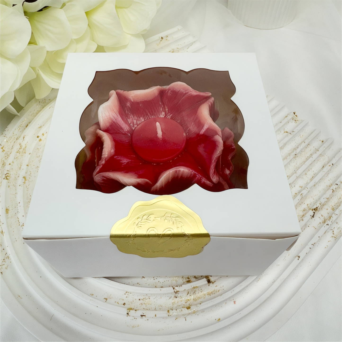 ine dunkelrote handgefertigte Blütenkerze in einer weißen Geschenkbox mit Sichtfenster und goldenem Siegel.