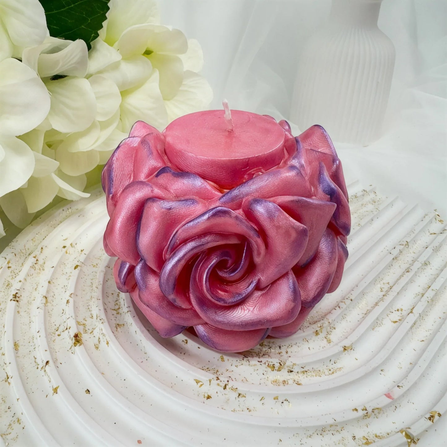Eine handgefertigte rosa Kerze in Form einer Rose mit schimmerndem Finish und violetten Details auf einem weißen gewellten Tablett.