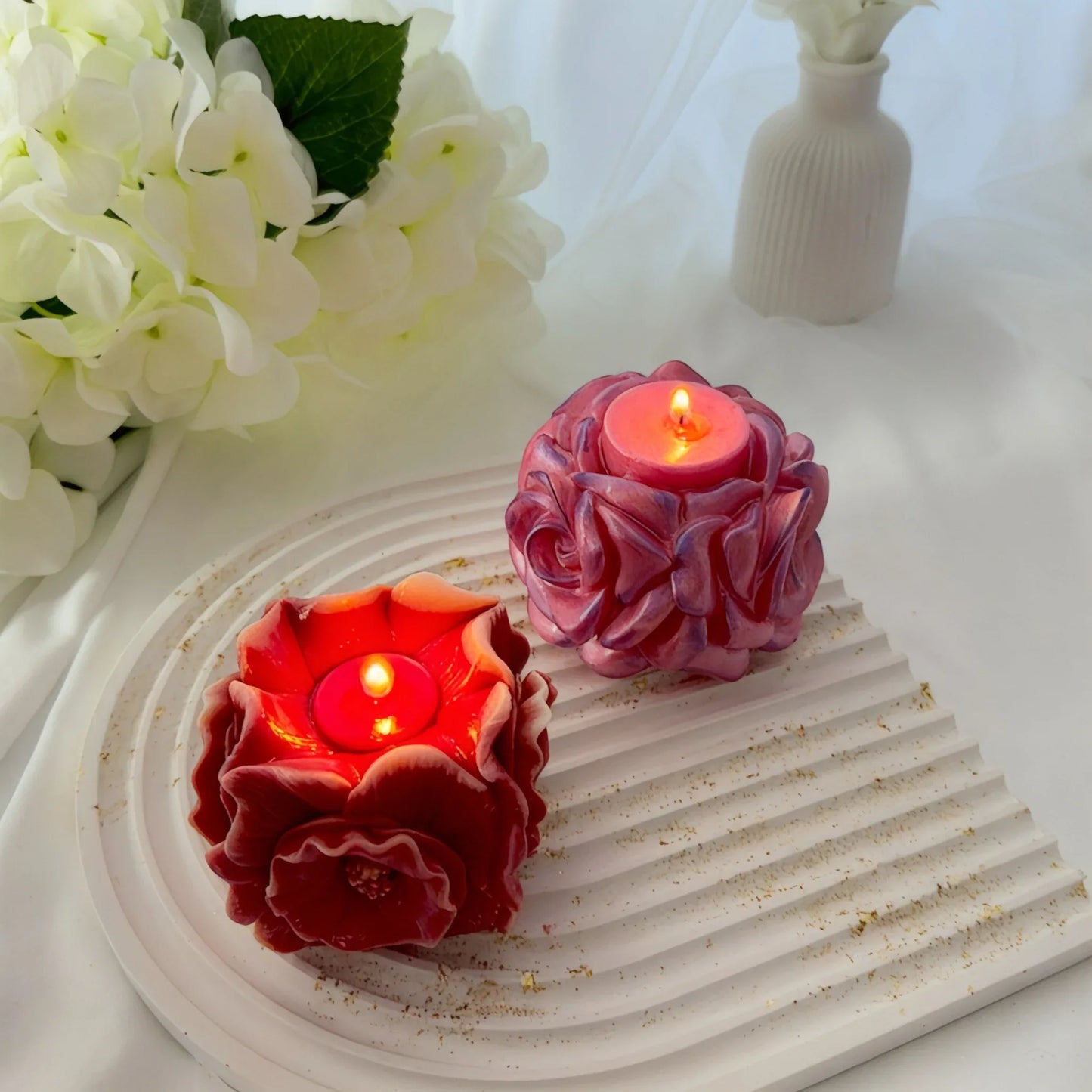 Zwei brennende handgefertigte Blütenkerzen – eine dunkelrote und eine schimmernde rosa Rose – auf einem weißen gewellten Tablett.