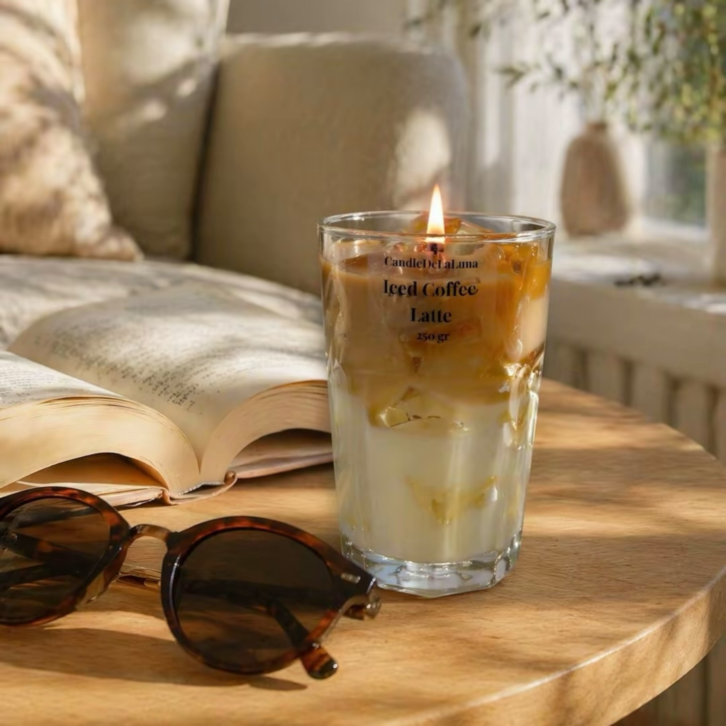 Eine brennende dekorative Kerze in Form eines Iced Coffee Latte steht in einem hohen, facettierten Glas auf einem Holztisch neben einer Sonnenbrille und einem aufgeschlagenen Buch, im Hintergrund ist ein gemütliches Wohnzimmer zu sehen.