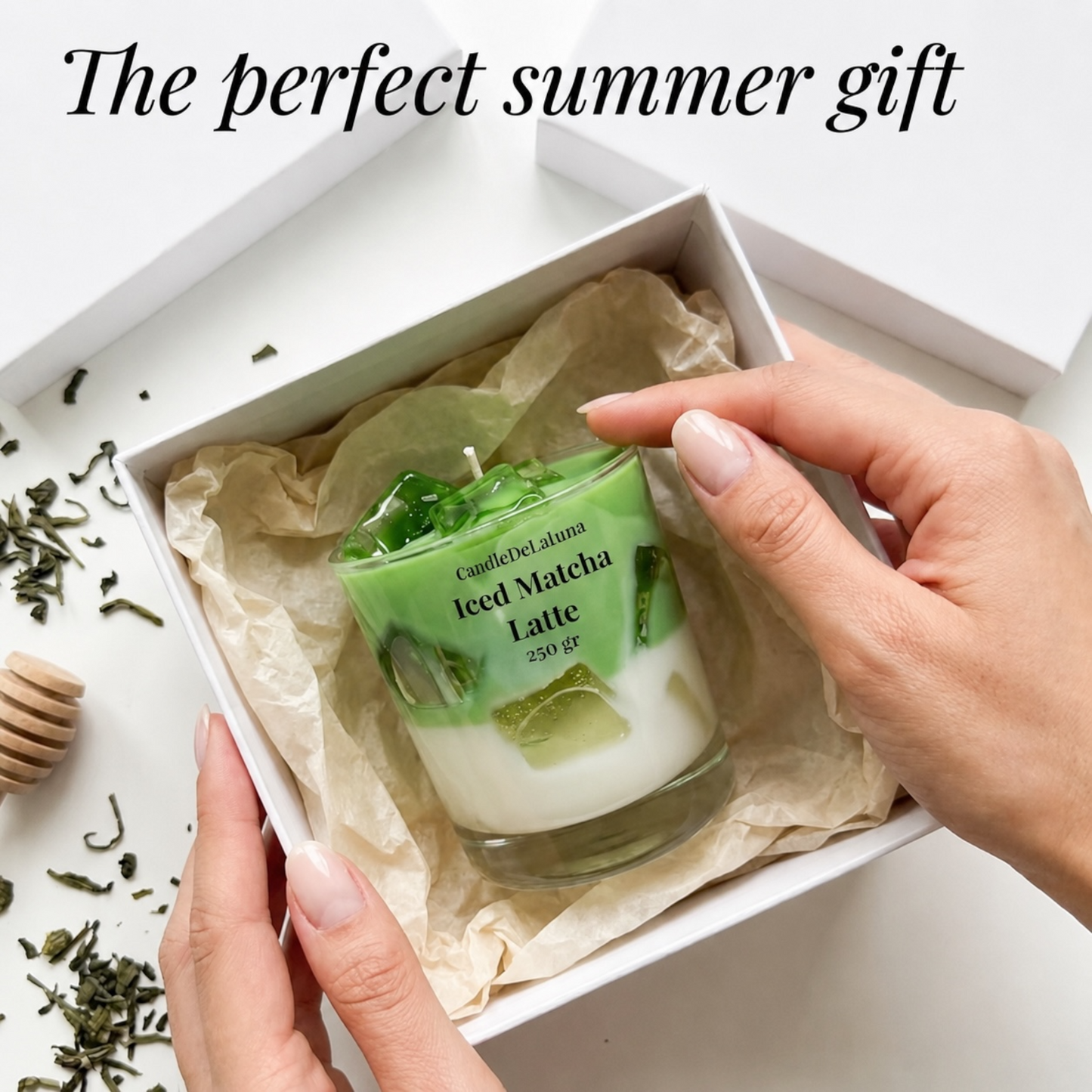 Eine Werbegrafik zeigt Hände, die eine dekorative Kerze in Form eines Iced Matcha Latte aus einer weißen Geschenkschachtel nehmen, ergänzt durch verstreute Teeblätter und den Text „The perfect summer gift“.