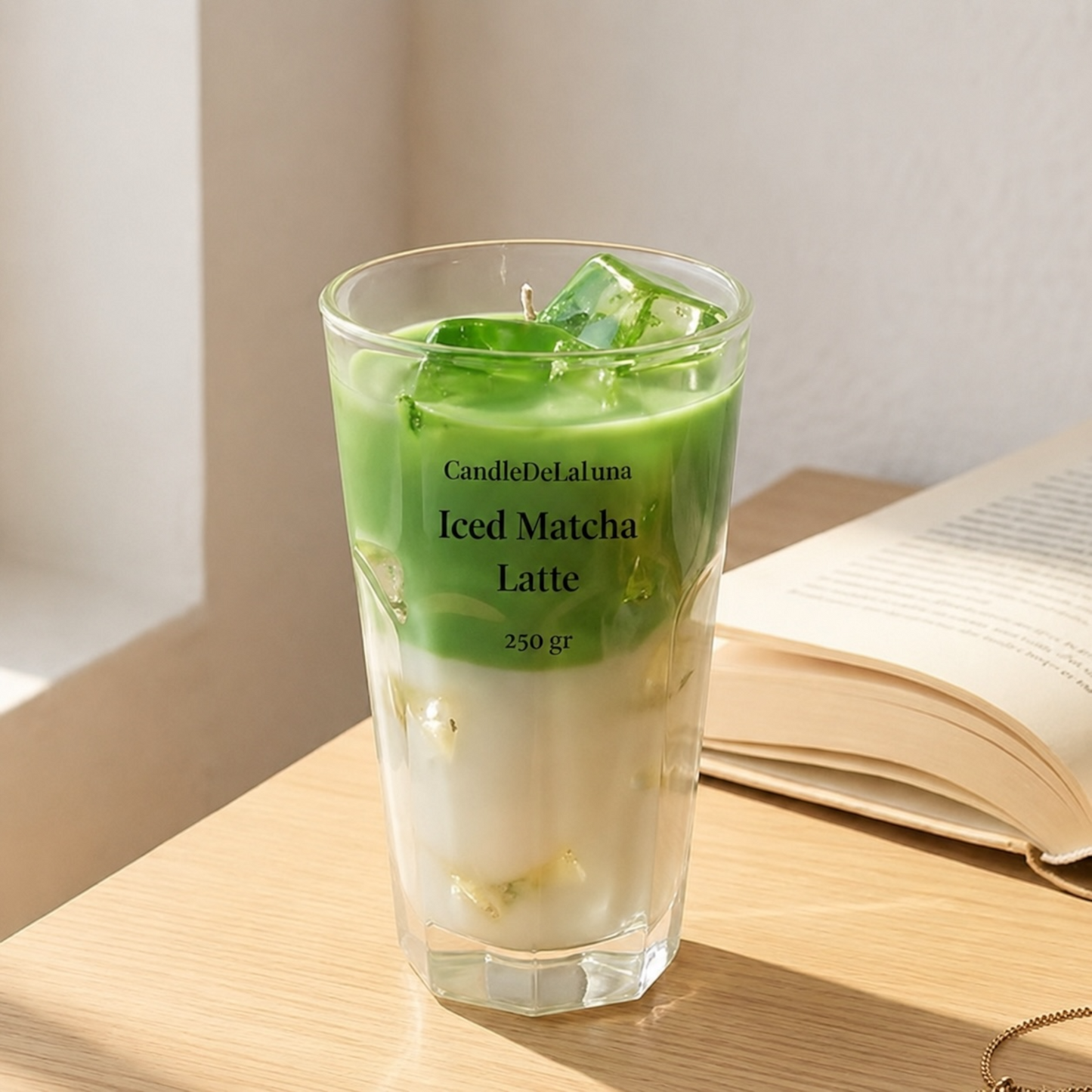 Eine dekorative Kerze in Form eines Iced Matcha Latte steht in einem hohen, facettierten Glas auf einem Holztisch neben einem aufgeschlagenen Buch, beleuchtet von warmem, natürlichem Licht.