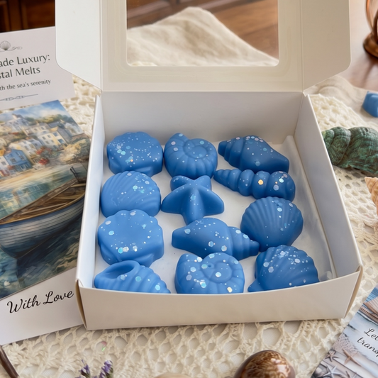 Eine quadratische weiße Geschenkbox mit Sichtfenster, gefüllt mit elf blauen, mit Glitzer verzierten Wachsschmelzen (Wax Melts) in verschiedenen maritimen Formen wie Seesternen und Muscheln, platziert auf einem hellen Spitzendeckchen neben einer Postkarte mit maritimem Motiv.