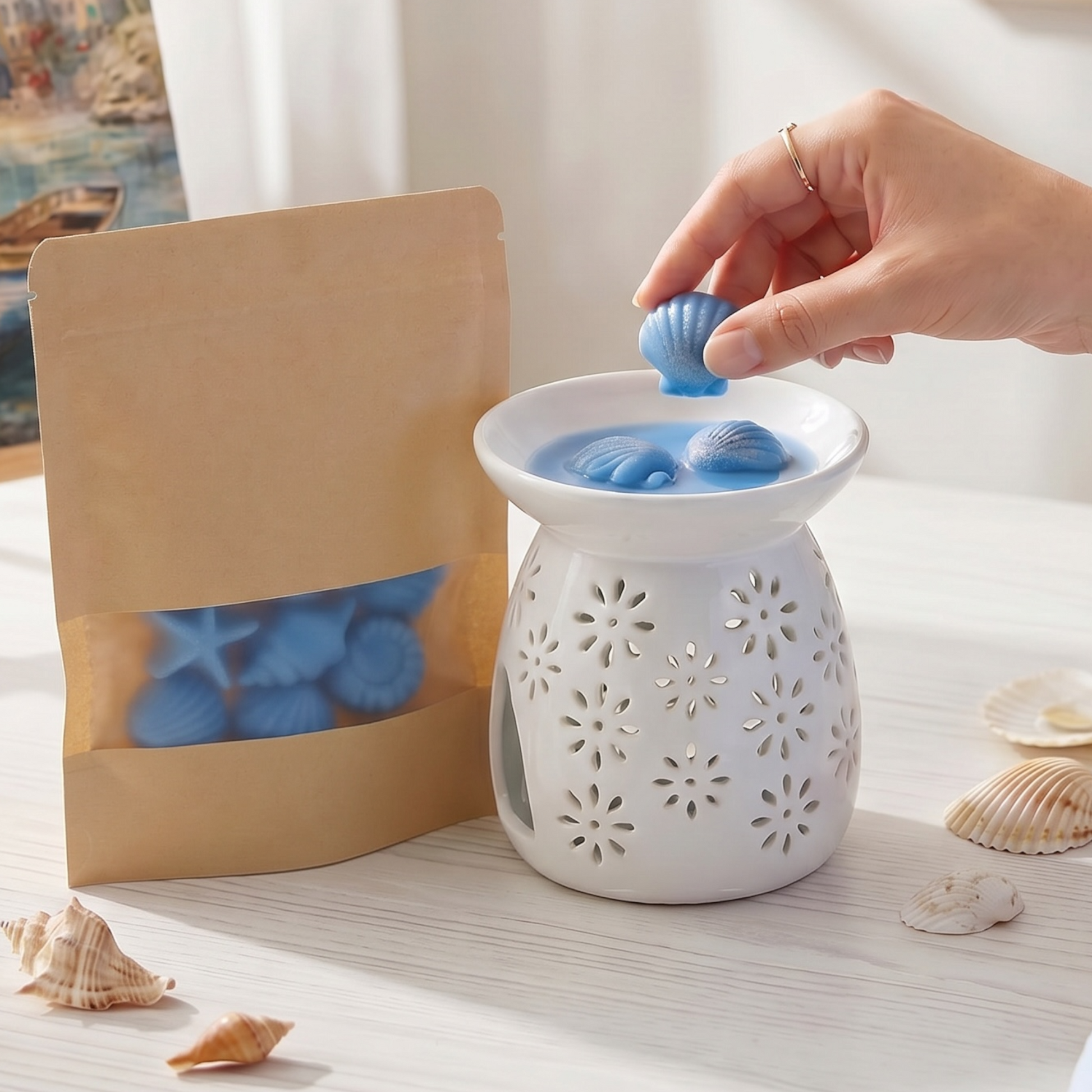 Eine Hand legt ein blaues, muschelförmiges Wachsschmelzstück (Wax Melt) in eine weiße Keramik-Aromalampe mit Blumenmuster, platziert auf einem hellen Tisch neben einer Kraftpapiertüte mit Sichtfenster und echten Seemuscheln.