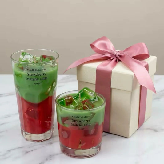 Zwei dekorative Kerzen in Form eines Strawberry Matcha Latte mit einer grünen oberen Schicht und einer roten unteren Schicht stehen neben einer beigen Geschenkschachtel, die mit einer großen rosa Satinschleife verziert ist, auf einer hellen Marmoroberfläche.
