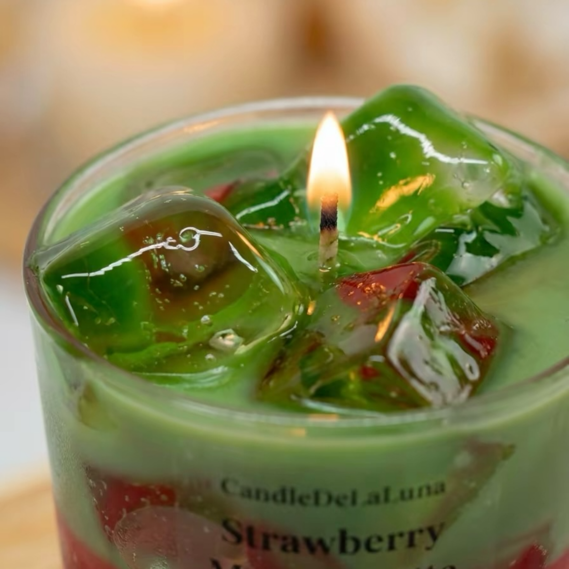 Eine brennende dekorative Kerze in Form eines Strawberry Matcha Latte mit einer grünen Oberfläche und transparenten Wachs-Eiswürfeln wird in einer Nahaufnahme gezeigt.