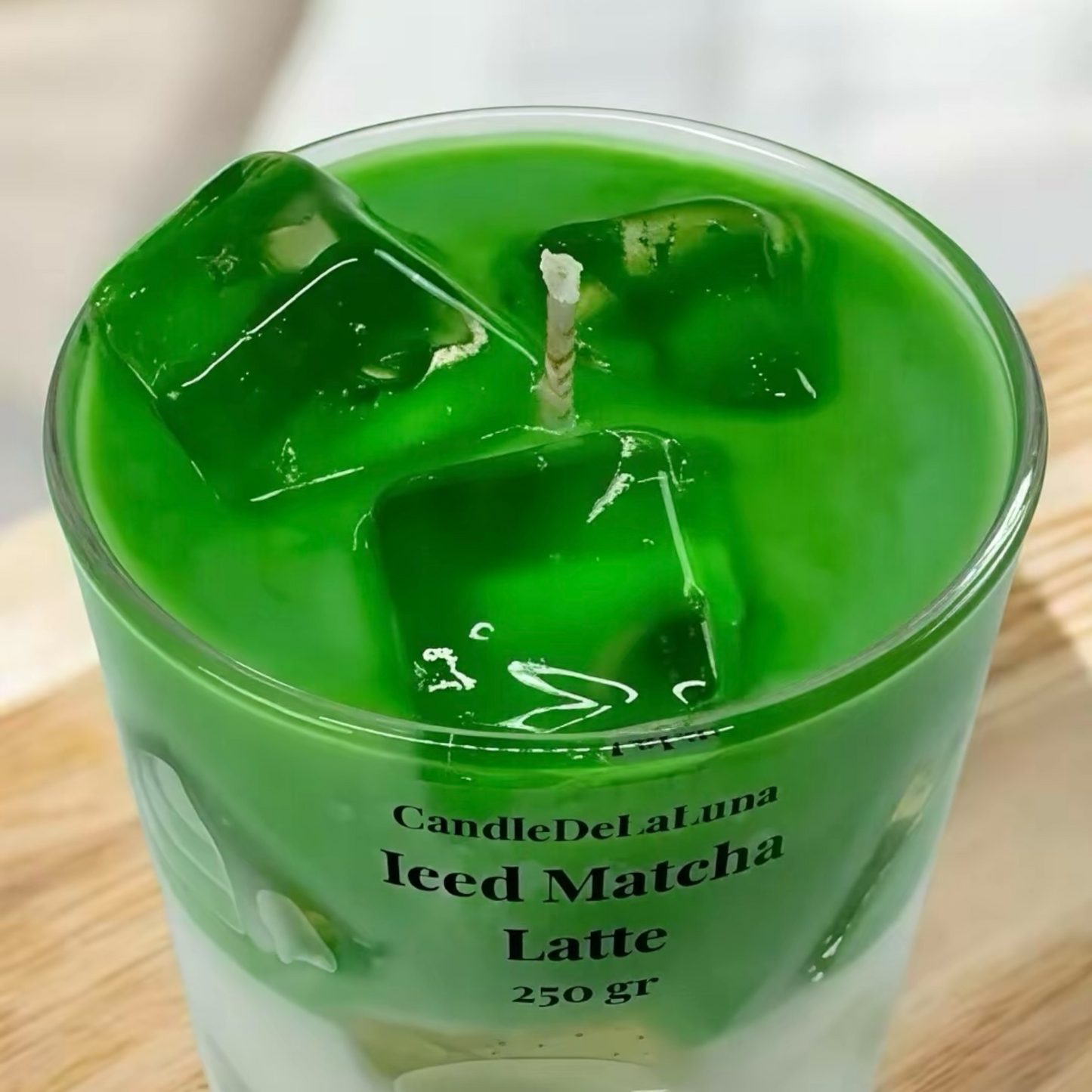 Eine dekorative Kerze in Form eines Iced Matcha Latte mit einer intensiv grünen Oberfläche und transparenten Wachs-Eiswürfeln wird in einer Nahaufnahme von oben auf einer Holzoberfläche gezeigt.