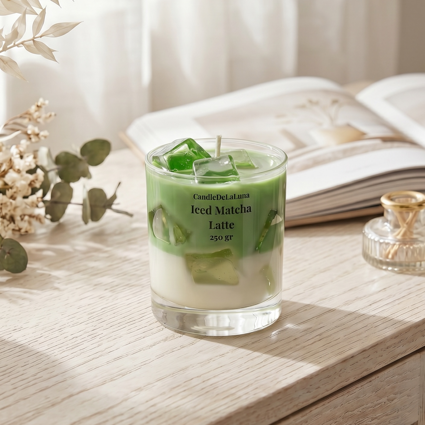 Eine dekorative Kerze in Form eines Iced Matcha Latte steht auf einer hellen Holzoberfläche neben einem geöffneten Buch, einem kleinen Flakon und Trockenblumen, umgeben von sanftem, natürlichem Licht.