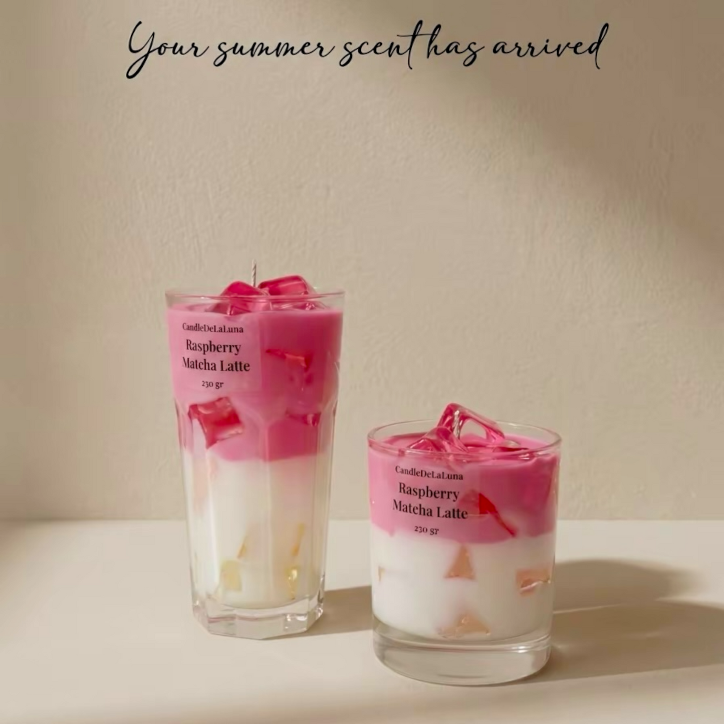 Eine Werbegrafik mit zwei dekorativen Kerzen in Form eines Raspberry Matcha Latte, bestehend aus einem hohen und einem niedrigen Glas vor hellem Hintergrund, ergänzt durch den geschwungenen Text „Your summer scent has arrived“.