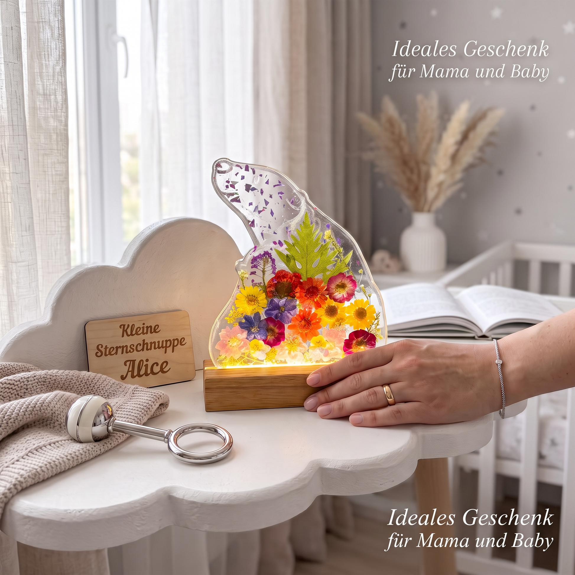Ein bärenförmiges Epoxidharz-Nachtlicht mit bunten Trockenblumen steht leuchtend auf einem weißen Kindertisch neben einer personalisierten Holzplakette und einer Babyrassel, während eine Hand den Sockel berührt, ergänzt durch den Werbetext „Ideales Geschenk für Mama und Baby“.