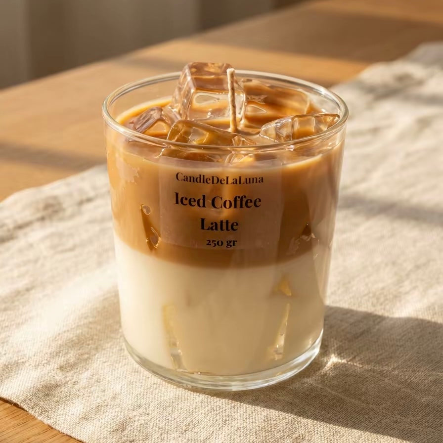 Eine dekorative Kerze in Form eines Iced Coffee Latte in einem niedrigen Glas steht auf einer hellen Tischdecke aus Leinen, beleuchtet von warmem, natürlichem Sonnenlicht.
